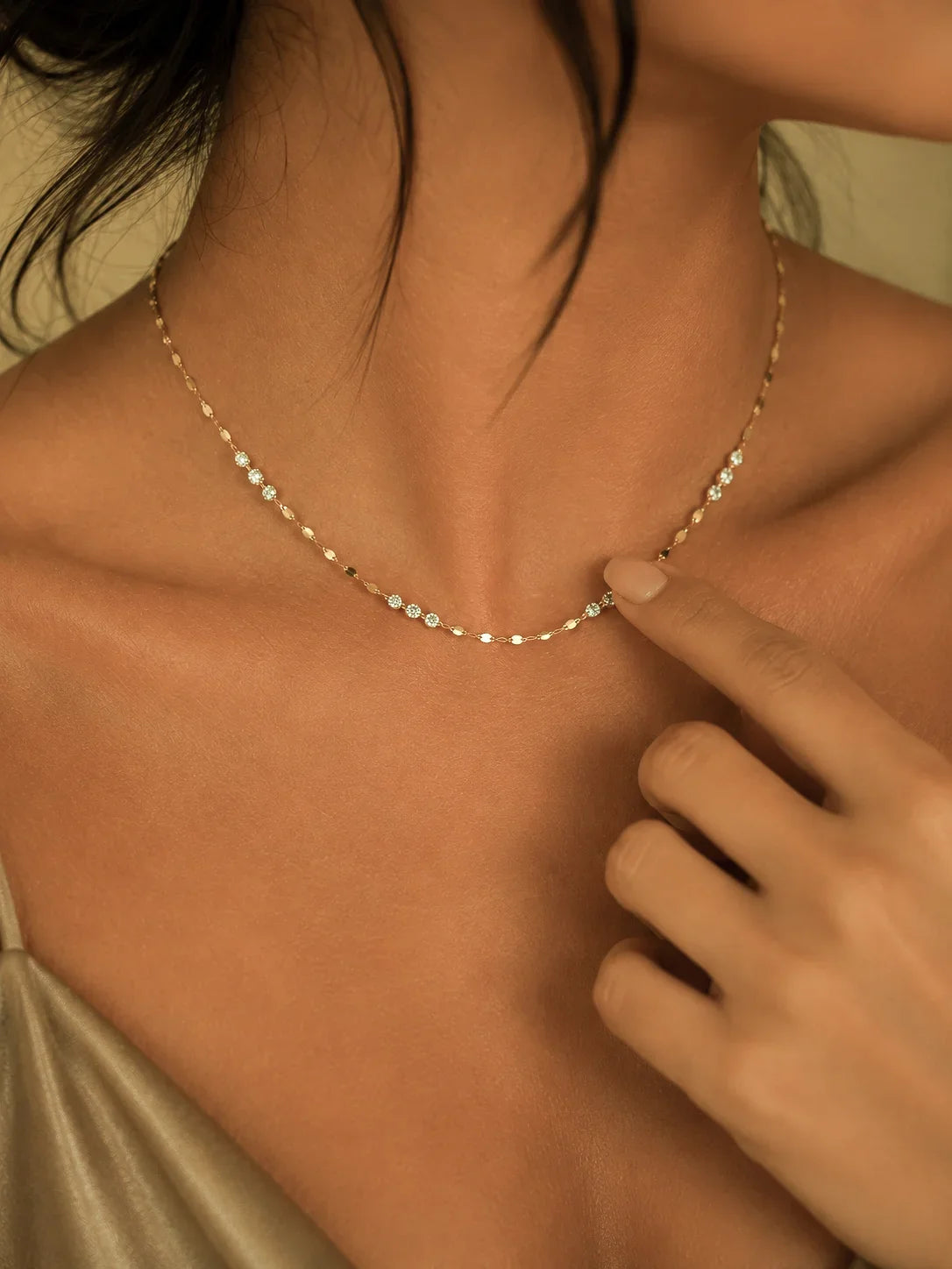 A delicate gold chain necklace with small clear stones worn by a model, with a finger pointing to a section of the necklace.