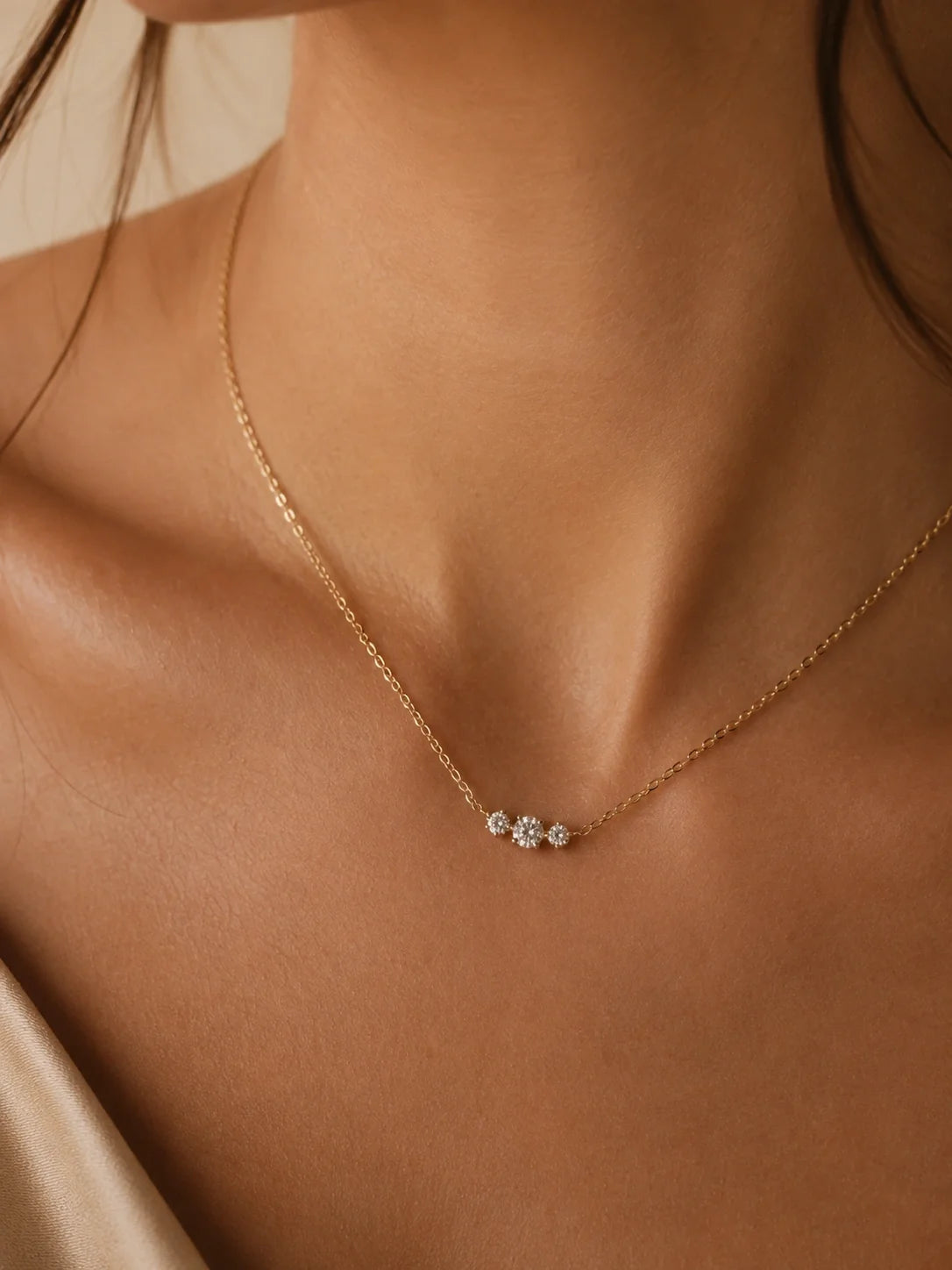 A delicate gold chain necklace with a small cluster of clear gemstones at the center, worn on a model's neck against a warm beige background.