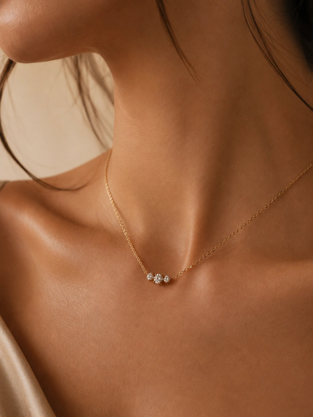 A delicate gold chain necklace with a small cluster of clear gemstones at the center, worn on a model's neck against a warm beige background.