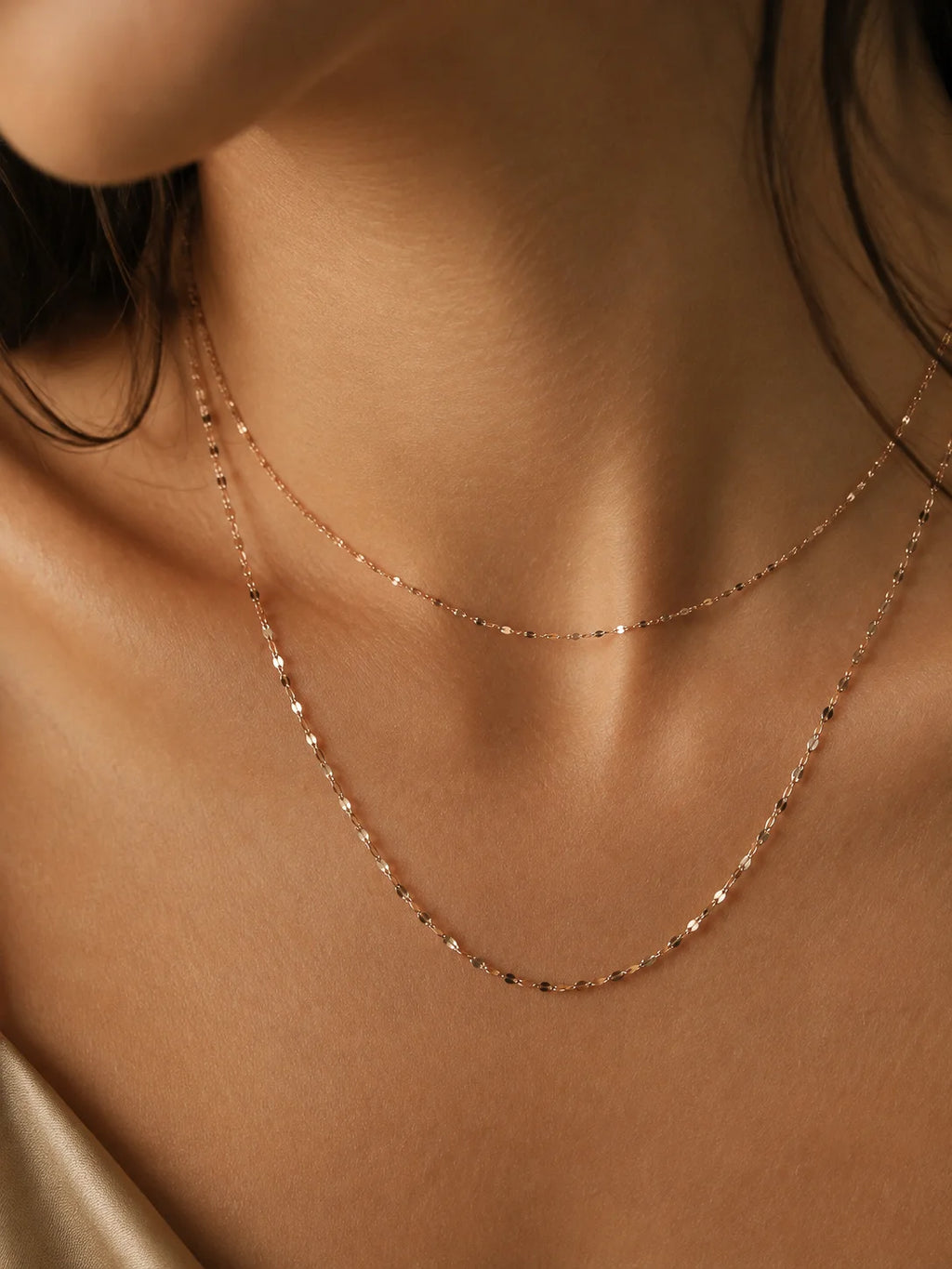 A close-up of a model's neck and upper chest wearing two delicate rose gold chain necklaces layered together.