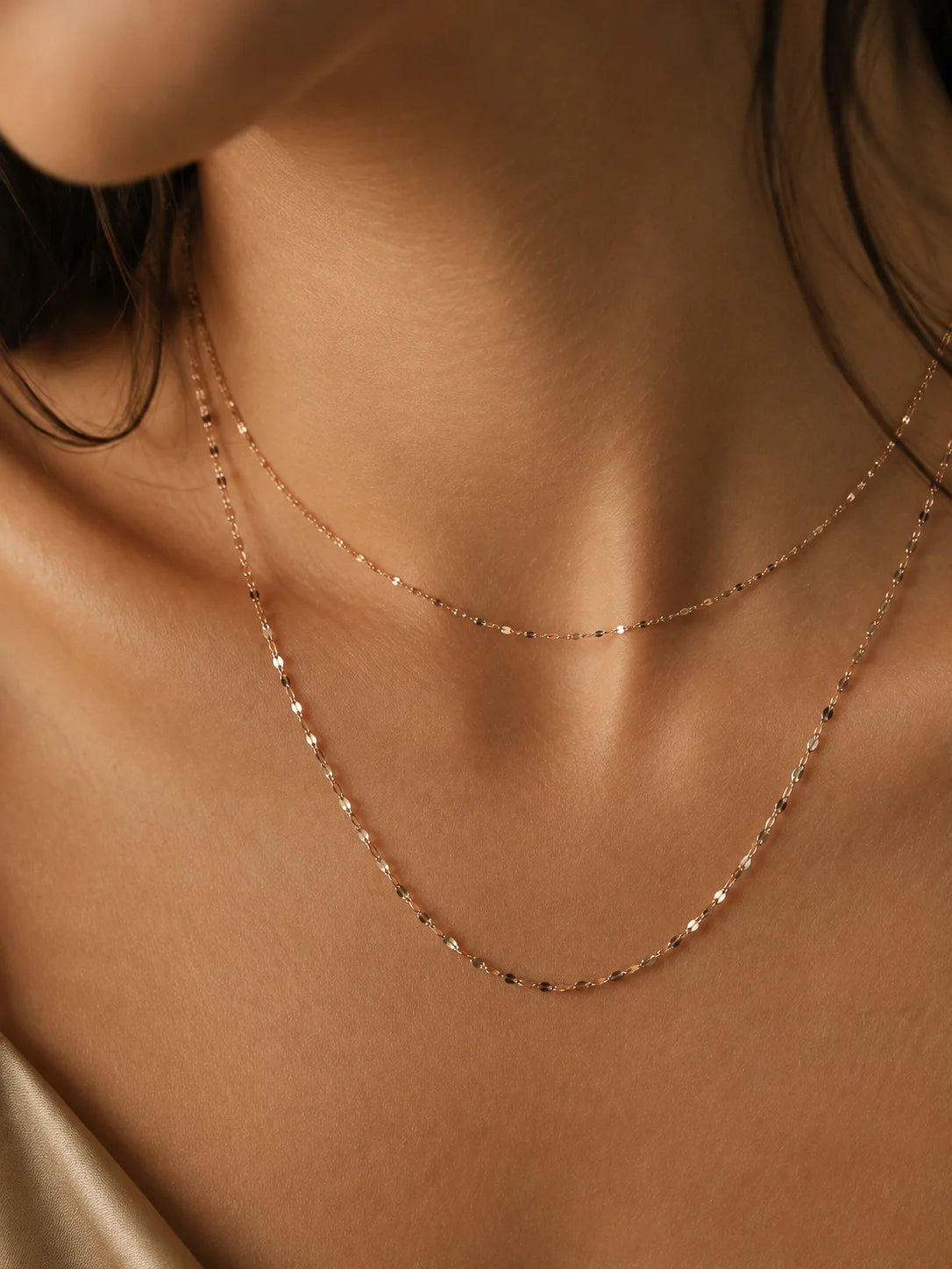 A close-up of a model's neck and upper chest wearing two delicate rose gold chain necklaces layered together.
