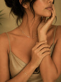 A woman wearing a delicate gold chain necklace and matching gold chain bracelet, styled with a beige satin camisole against a warm, neutral background.