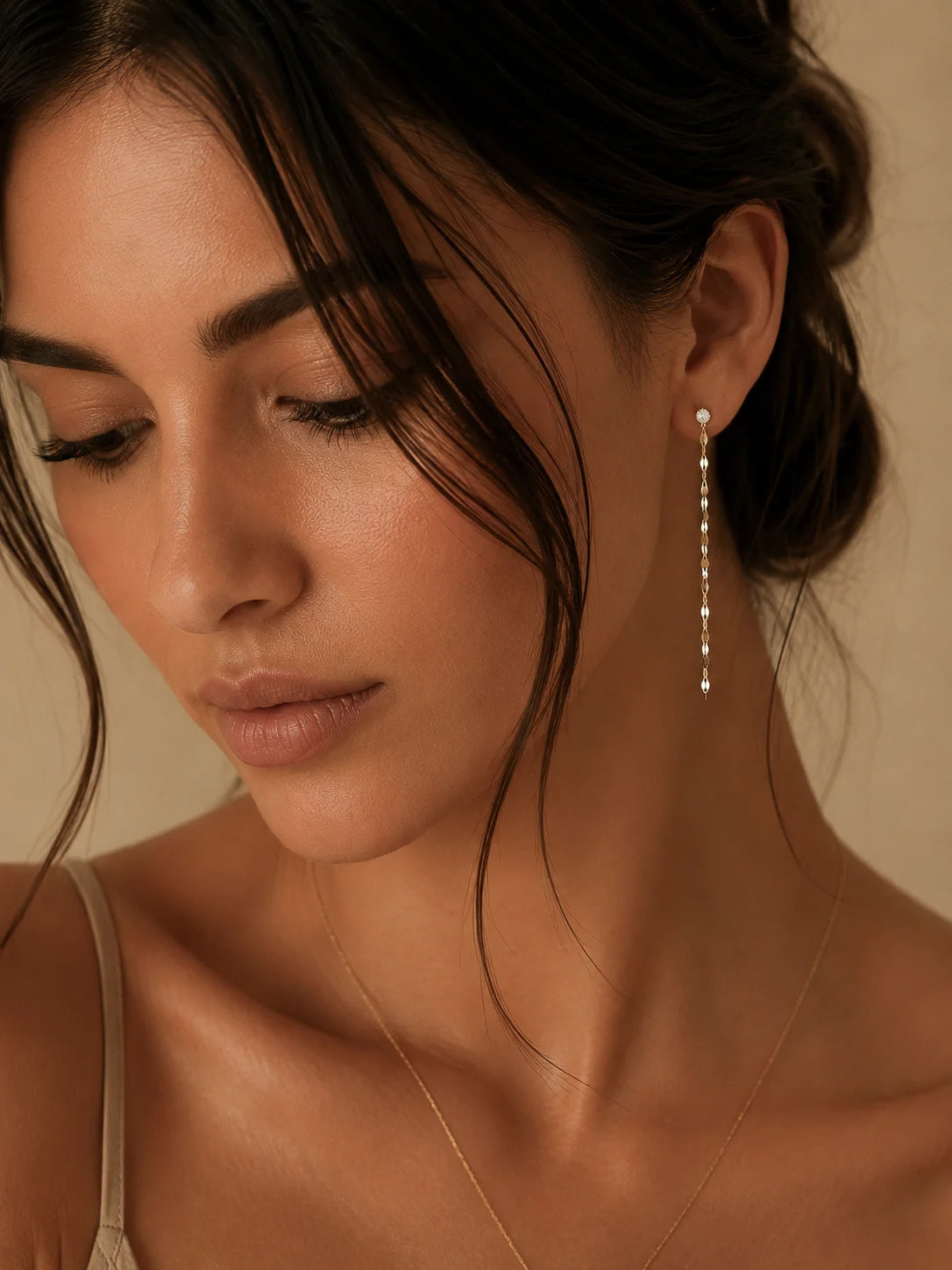 A close-up of a woman wearing a delicate silver dangle earring with small clear stones, set against a warm beige background.