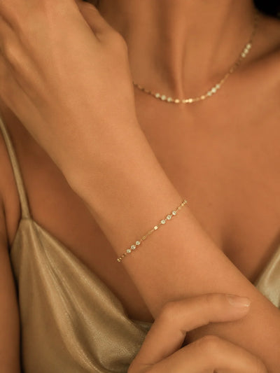 A delicate gold chain bracelet with small clear stones worn on a woman's wrist, shown in a warm-toned close-up with a matching necklace also visible.