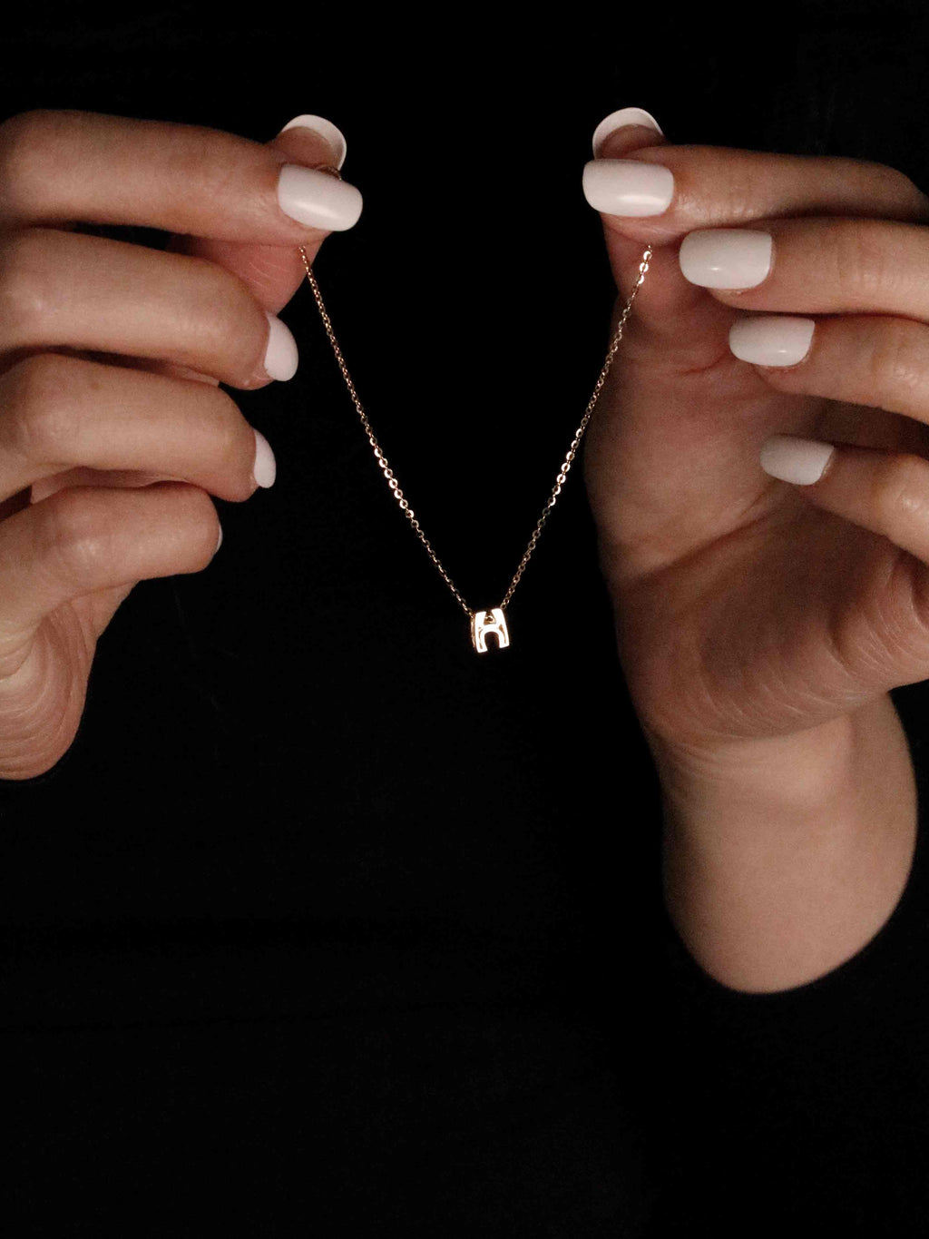 Hands holding a bracelet with a 'H' pendant against a black background
