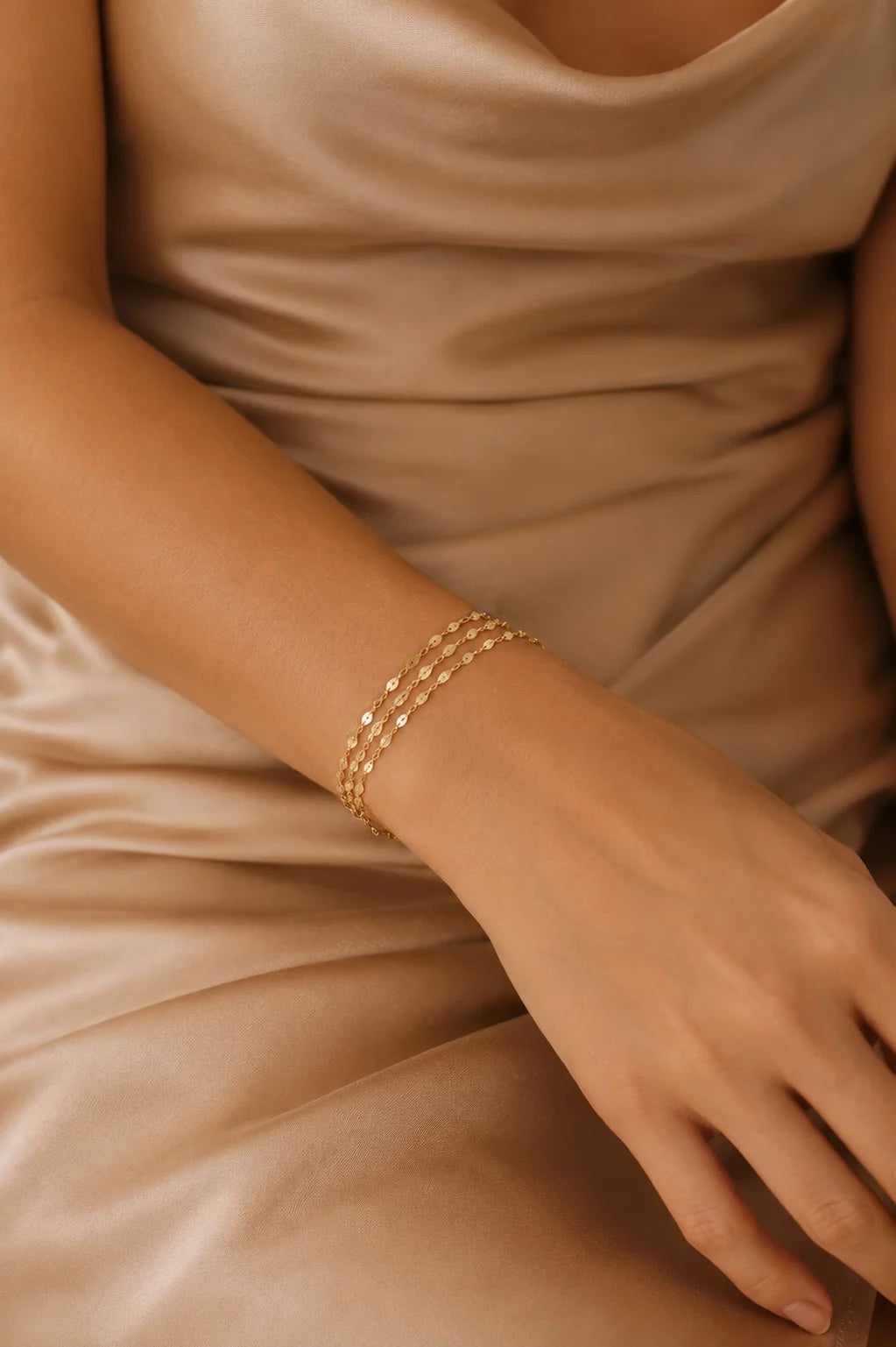 A close-up of a person wearing a delicate gold multi-strand chain bracelet on their wrist, dressed in a satin champagne-colored dress.