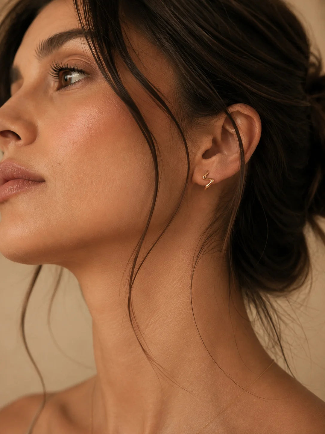 Close-up of a woman wearing a delicate gold crescent moon stud earring, shown in profile against a warm beige background.