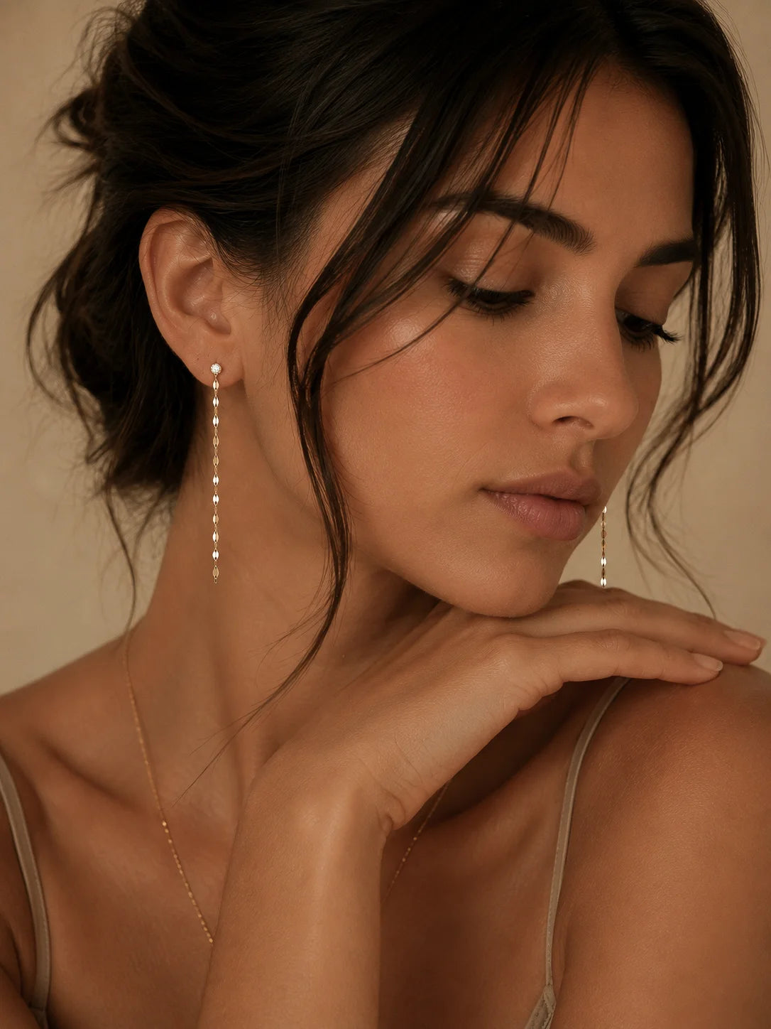 A close-up lifestyle shot of a woman wearing delicate gold dangle earrings with small white pearls, styled with a thin gold chain necklace against a warm beige background.