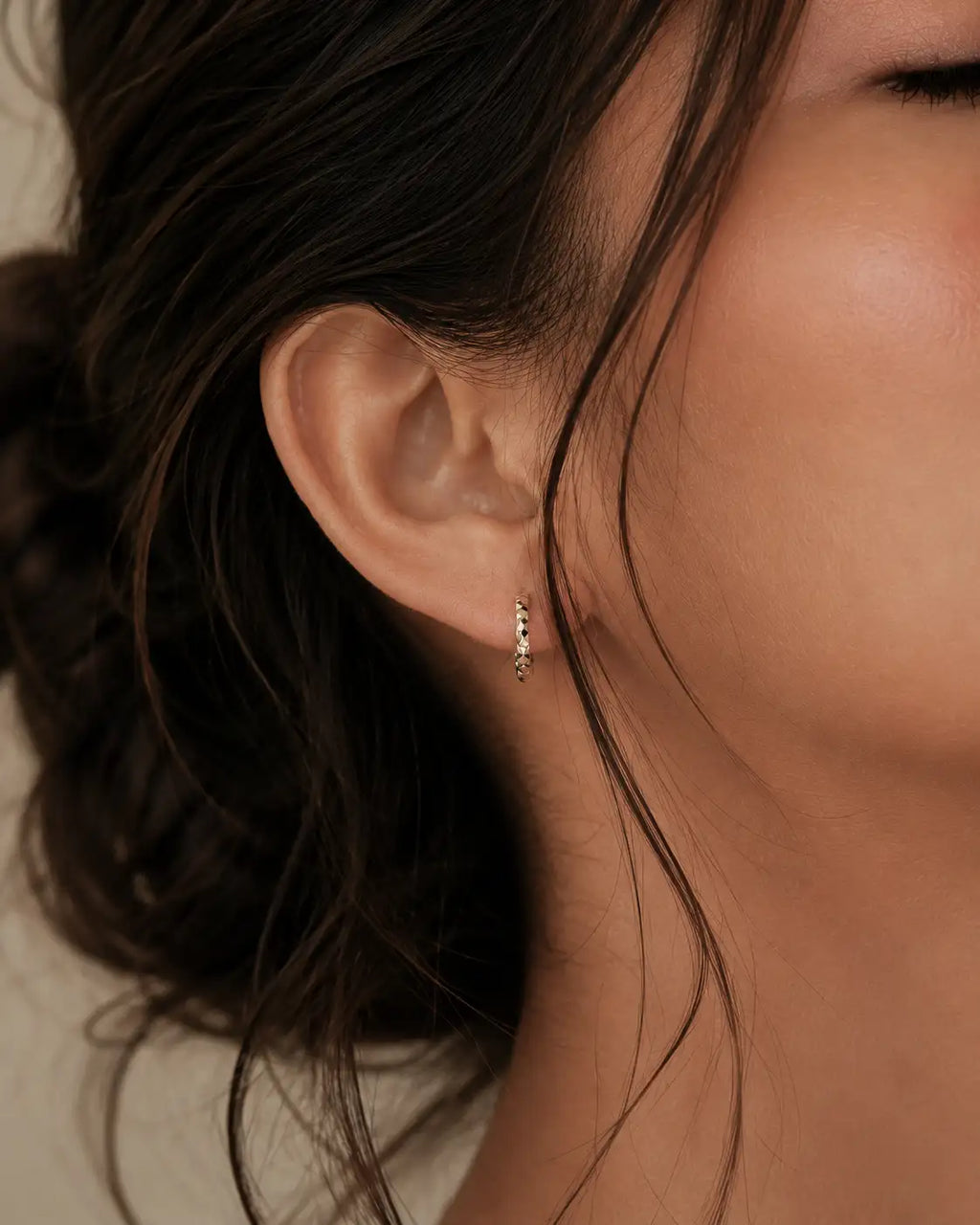 A close-up of a model's ear wearing a delicate gold huggie hoop earring with small clear stones.