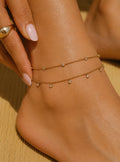 A close-up of a person's ankle and foot wearing a delicate double-layered gold anklet with small dangling bead details, with a hand touching the ankle.