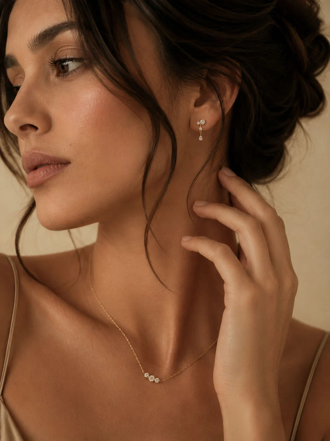 A close-up lifestyle shot of a woman wearing delicate gold jewelry, featuring a fine chain necklace with a small cluster of white stones and matching drop earrings.