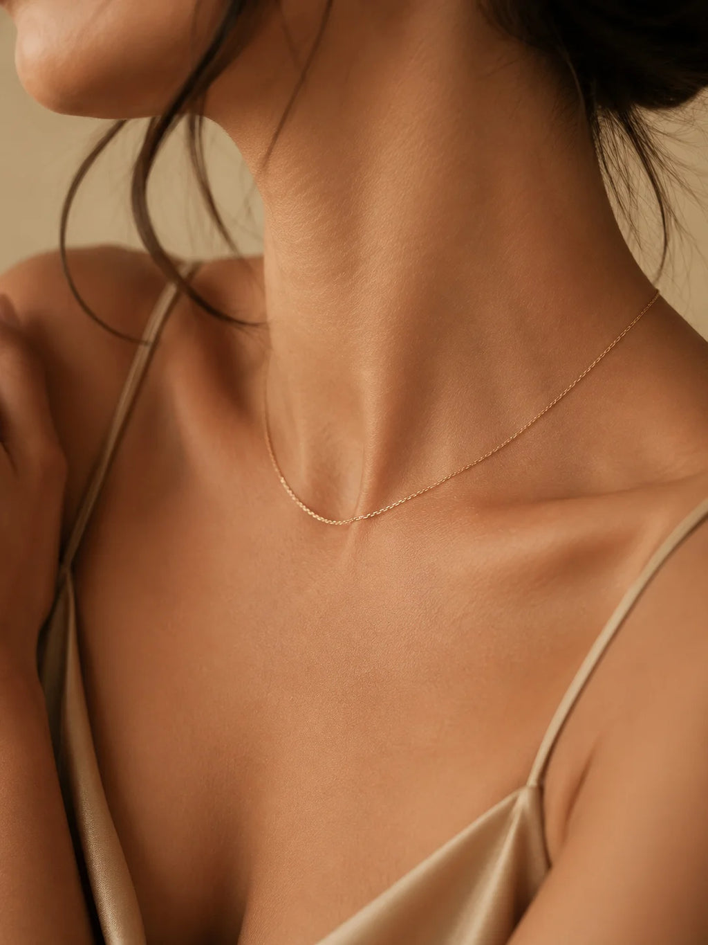 A delicate gold chain necklace worn by a model in a close-up shot, wearing a satin camisole against a warm beige background.