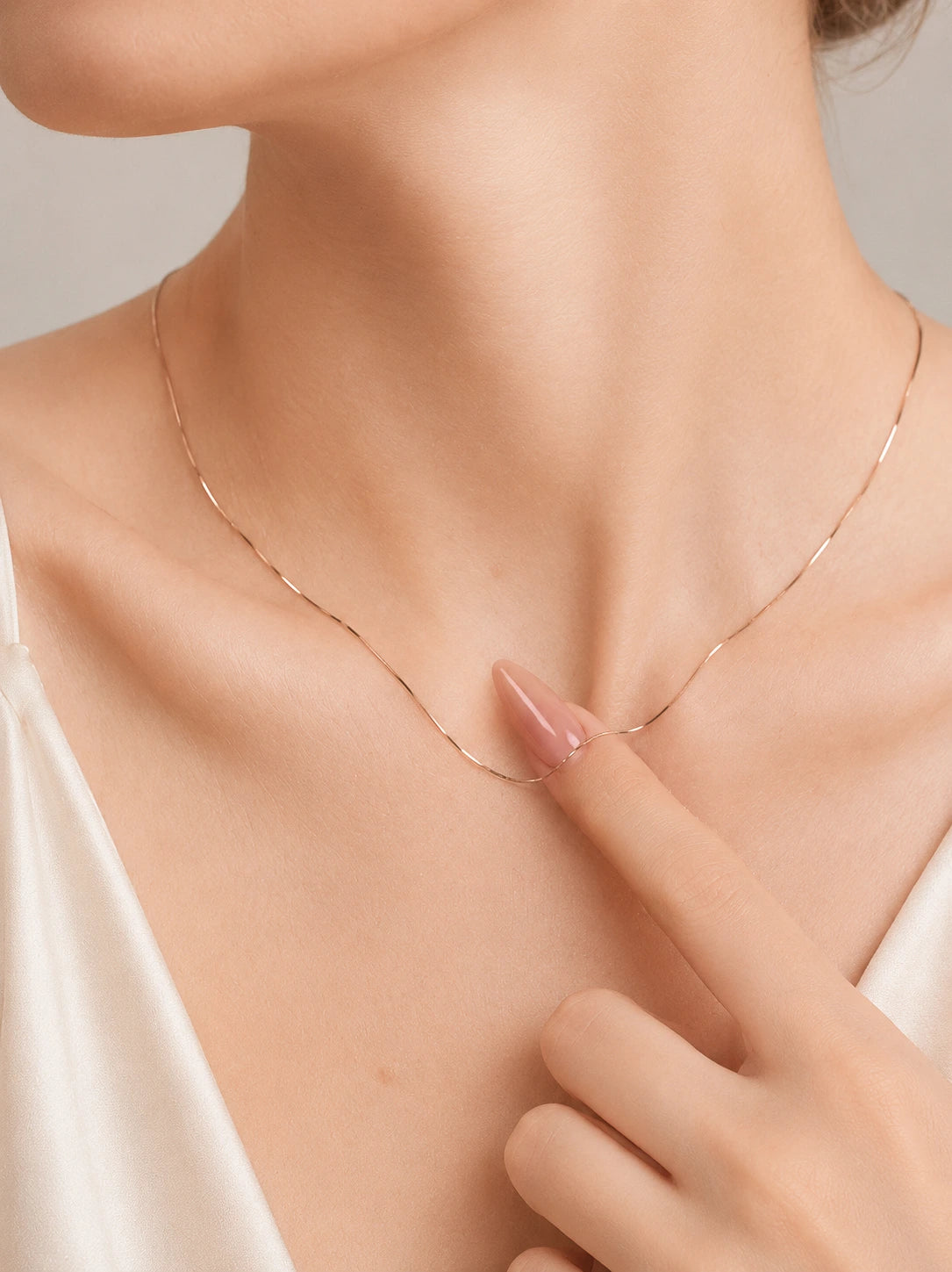 Close-up of 18K solid gold snake chain necklace on woman's collarbone, finger lifting the flat gold chain, styled with cream satin cami