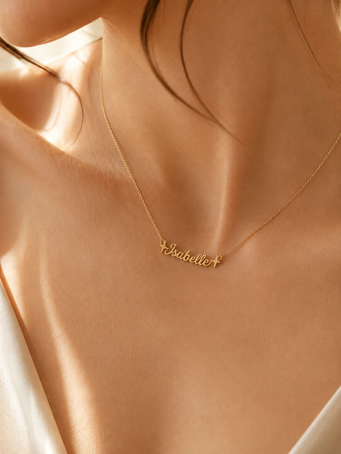 A close-up of a model's neck and upper chest wearing a delicate gold chain necklace with a personalized script nameplate reading 'Isabelle'.