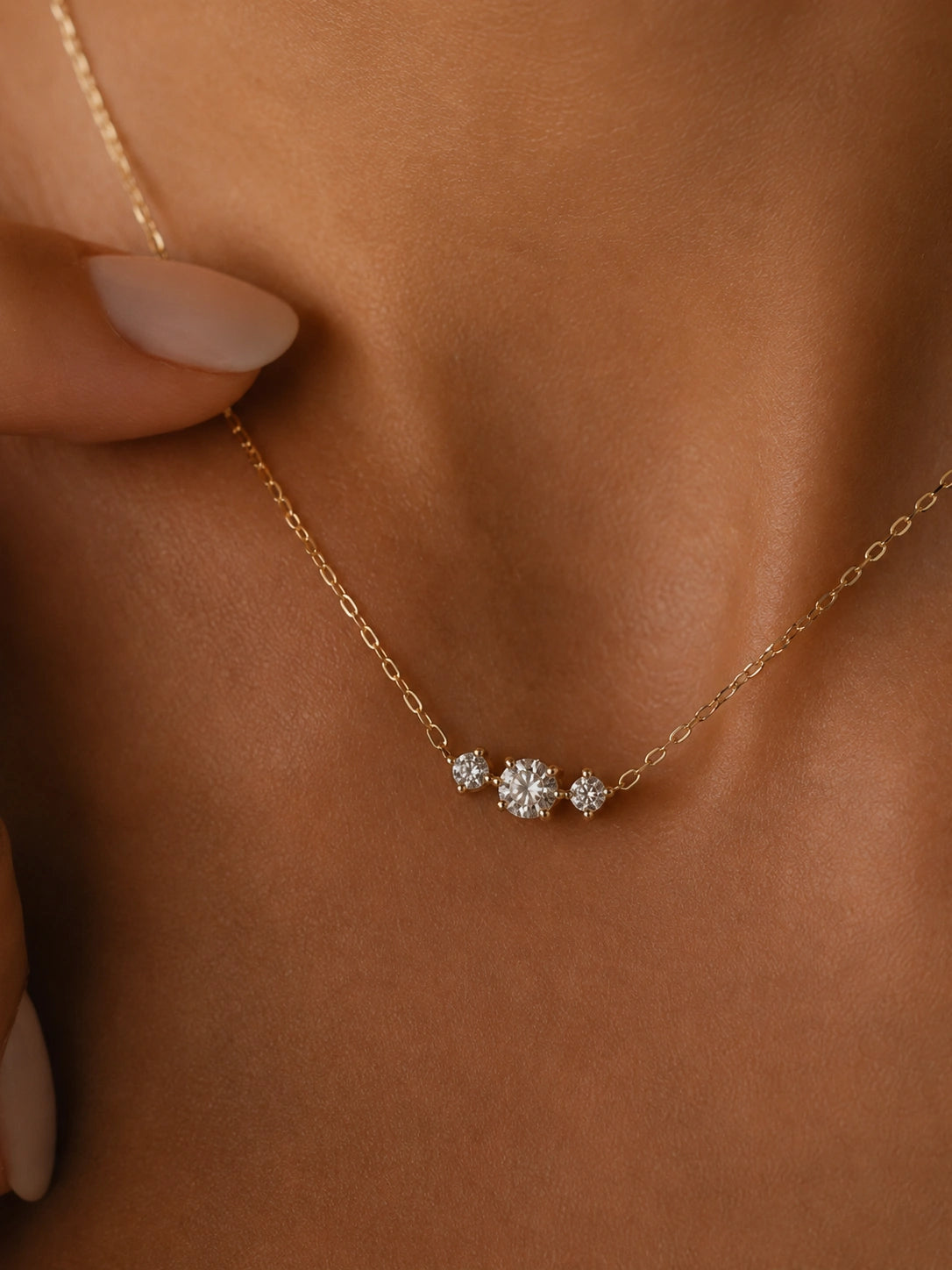 A delicate gold chain necklace with three small clear gemstone clusters, worn on a model's neck with a close-up of the skin and manicured nails visible.