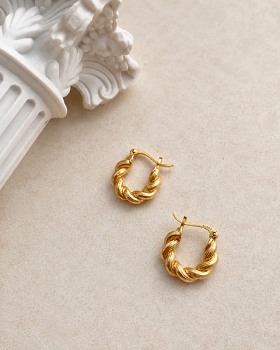 A pair of gold hoop earrings with a twisted rope texture, displayed on a light beige textured surface next to a white decorative classical column prop.