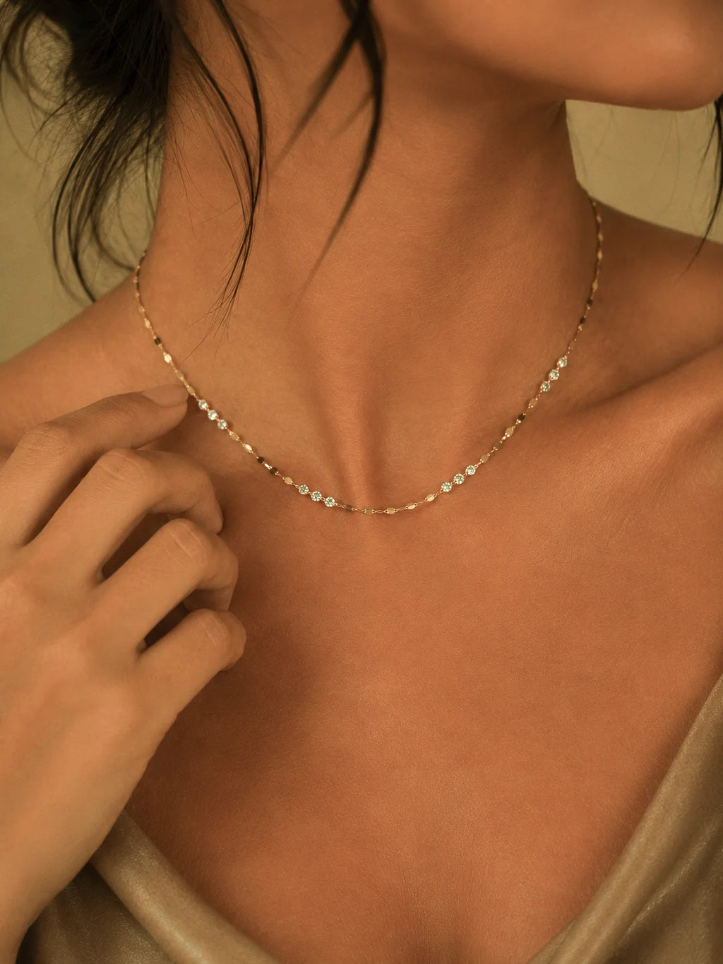 A delicate gold chain necklace with small clear stones worn by a model. The shot is a close-up of the model's neck, chest, and hand adjusting the necklace, with warm, soft lighting.