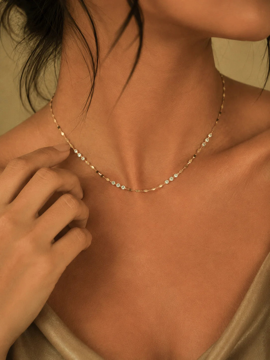 A delicate gold chain necklace with small clear stones worn by a model. The shot is a close-up of the model's neck, chest, and hand adjusting the necklace, with warm, soft lighting.