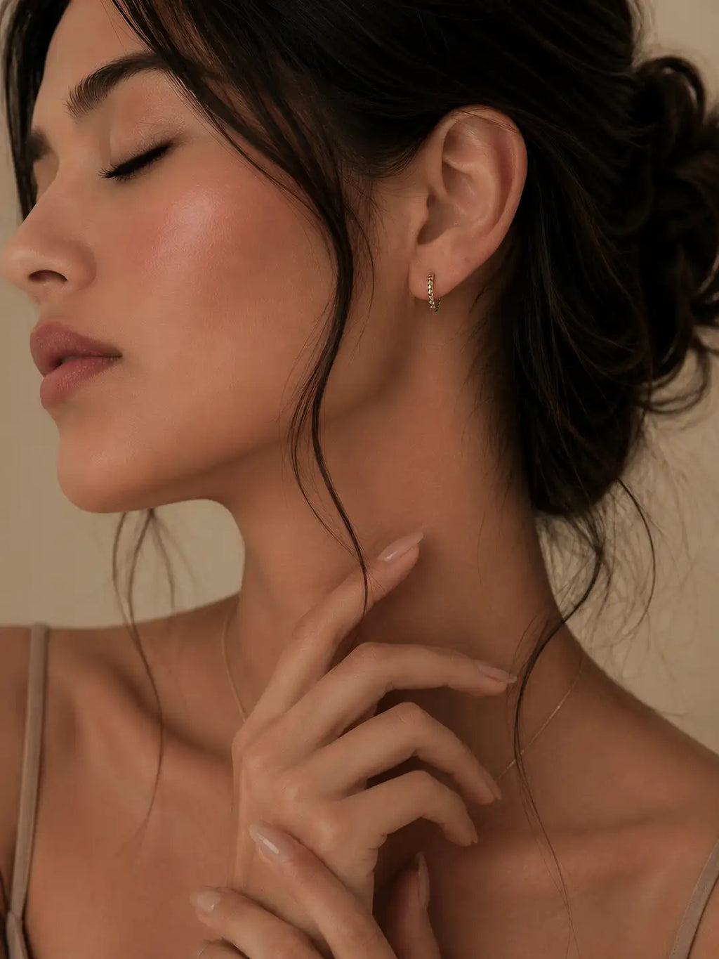 A close-up lifestyle shot of a woman with her eyes closed, wearing a delicate gold hoop earring with a small clear gemstone. Her hand gently rests near her neck, and the background is a soft, warm beige.