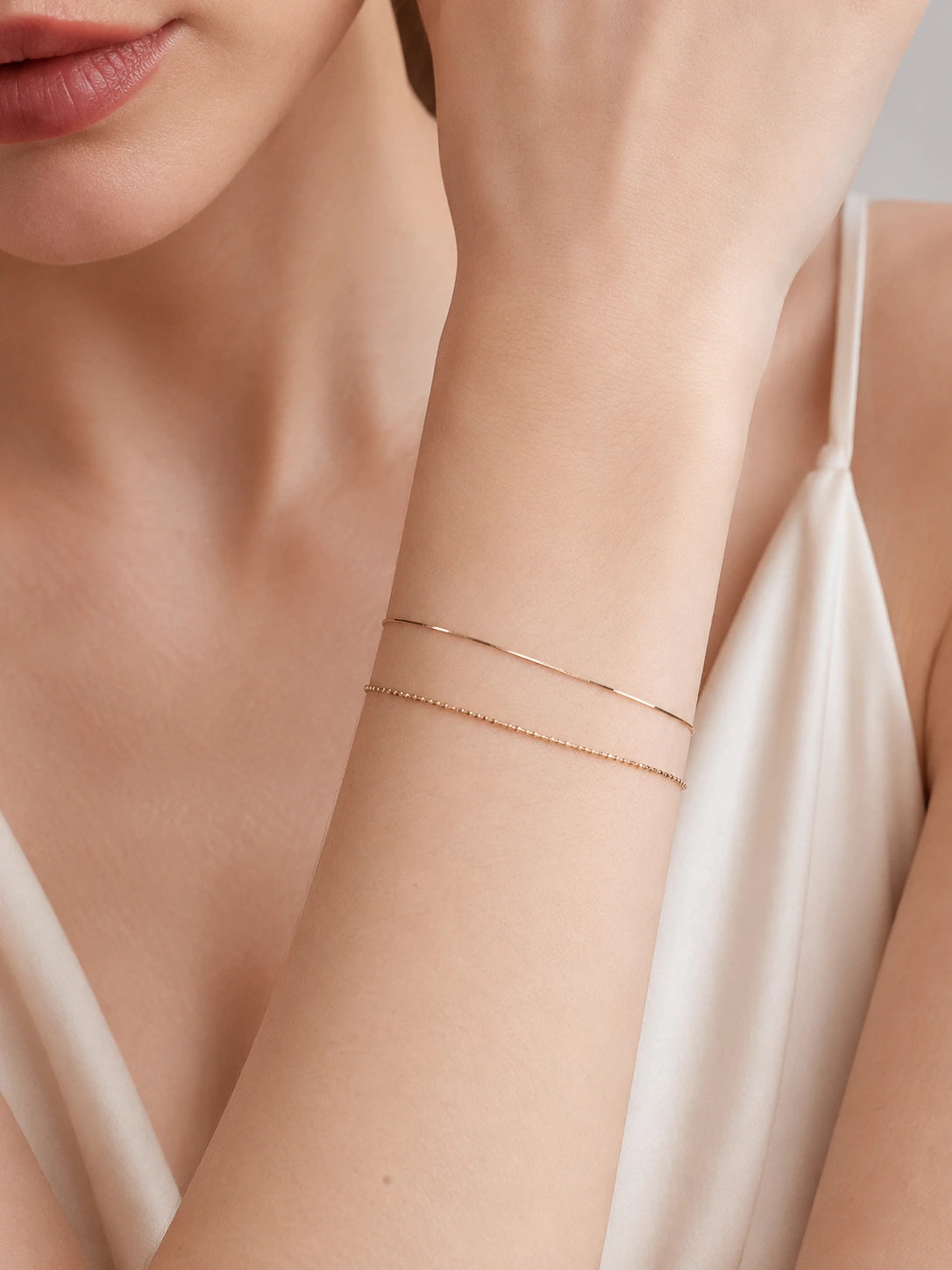 Woman wearing 18K solid gold snake chain bracelet on wrist and matching thin gold snake chain necklace, styled with ivory silk cami