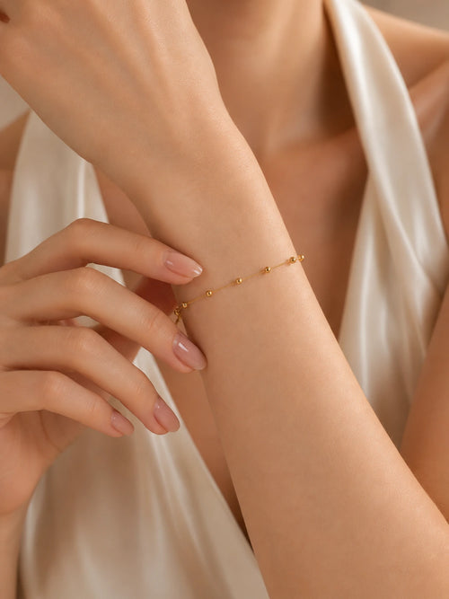 Dainty 18K solid gold satellite bead bracelet on wrist, hand detail shot showing gold ball stations on fine chain, cream satin background