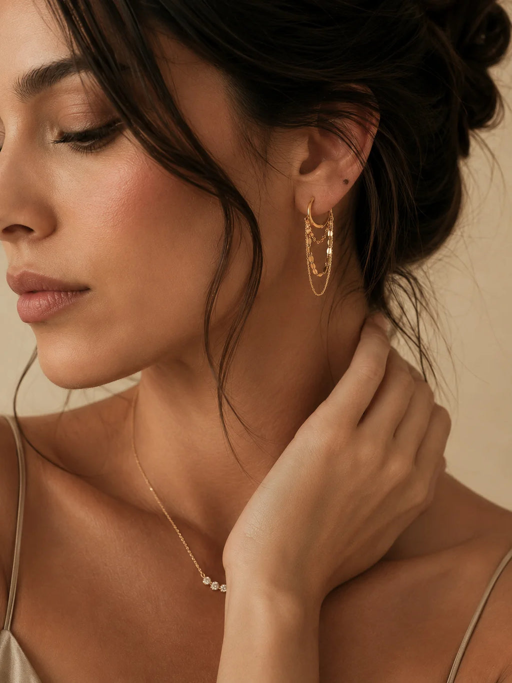 A close-up lifestyle shot of a woman wearing a delicate gold chain necklace and matching gold hoop earrings, with her hand gently touching her neck against a warm beige background.
