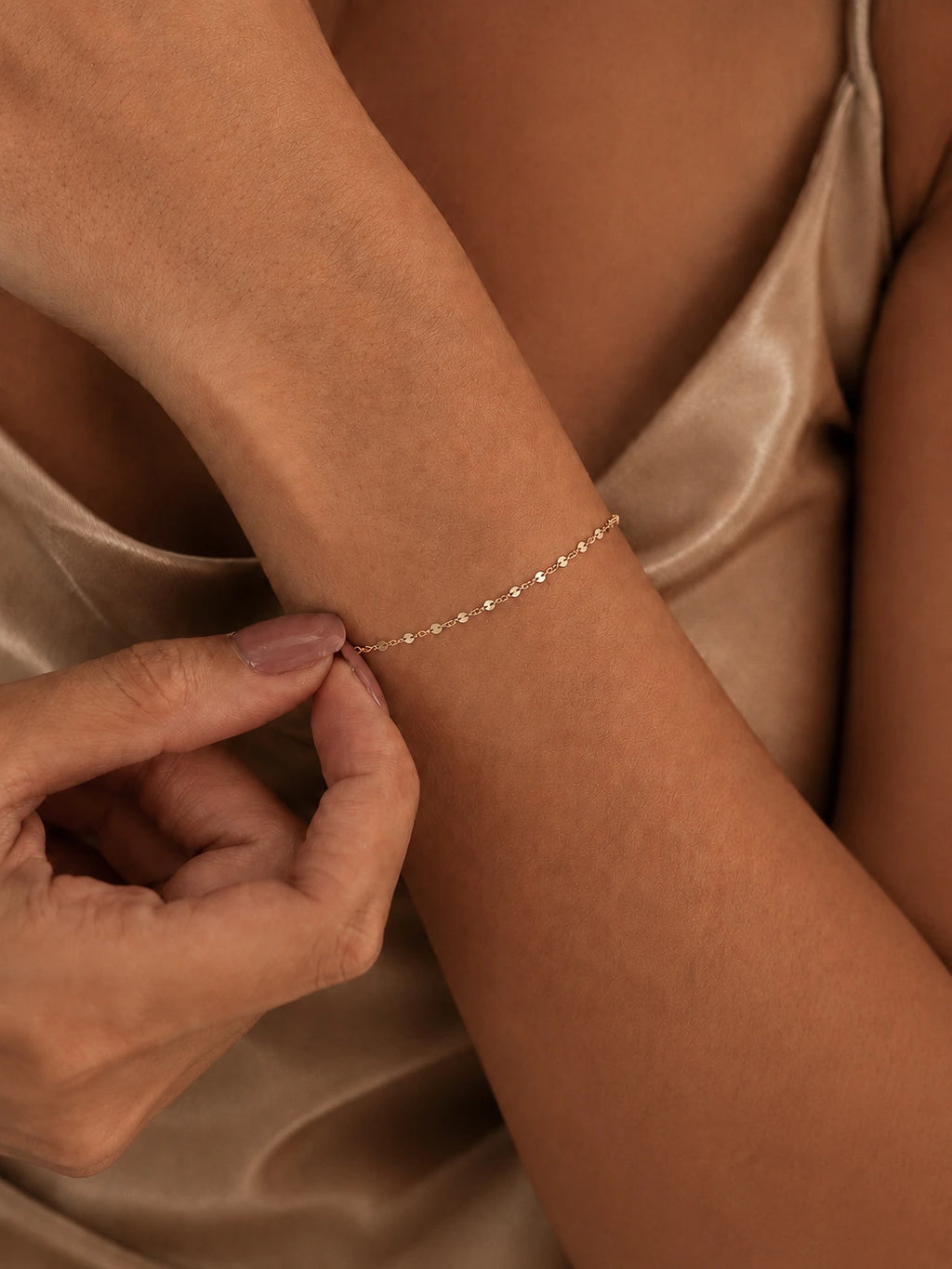 A delicate gold chain bracelet worn on a woman's wrist, shown in a close-up lifestyle shot with her hand adjusting it. She is wearing a champagne-colored satin top.