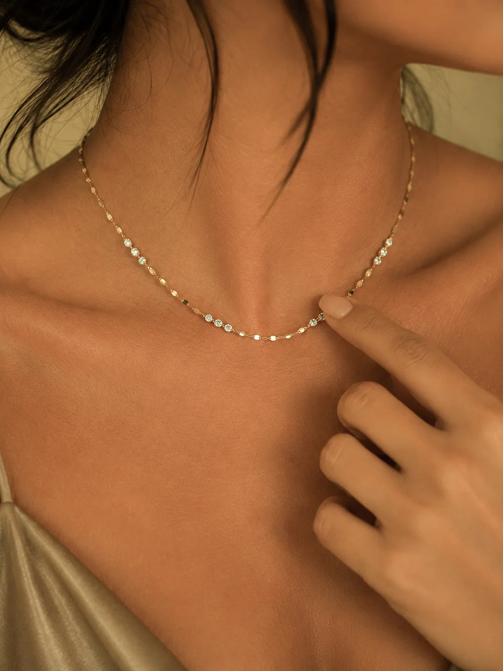 A delicate gold chain necklace with small clear stones worn by a model, with a finger pointing to a section of the necklace.