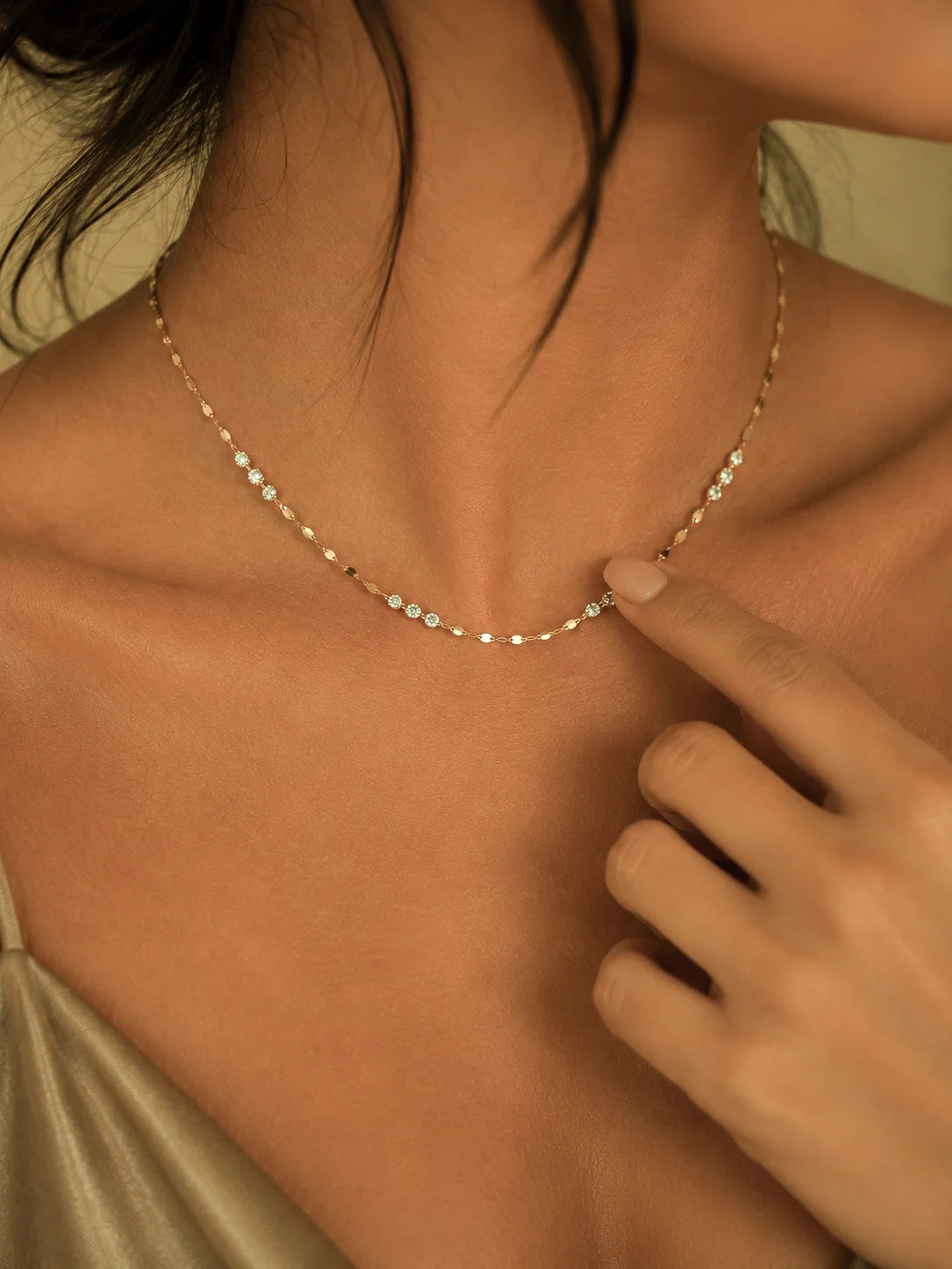 A delicate gold chain necklace with small clear stones worn by a model, with a finger pointing to a section of the necklace.