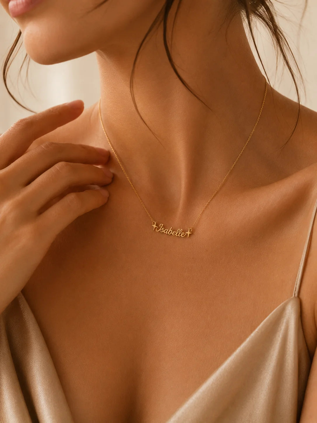 A close-up lifestyle shot of a model wearing a delicate gold script necklace with the name 'Isabel' and a small cross pendant, styled with a champagne satin top.