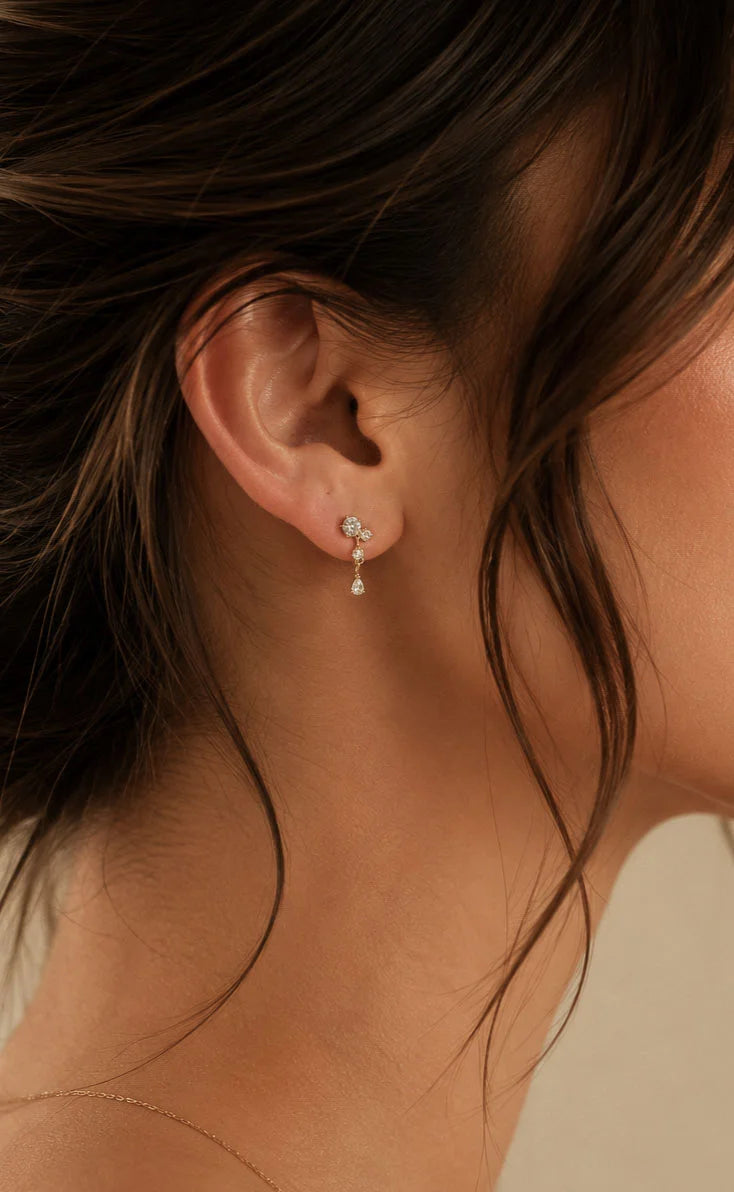 A close-up side profile of a woman's ear wearing a delicate silver dangle earring with a small clear gemstone and a tiny teardrop charm.