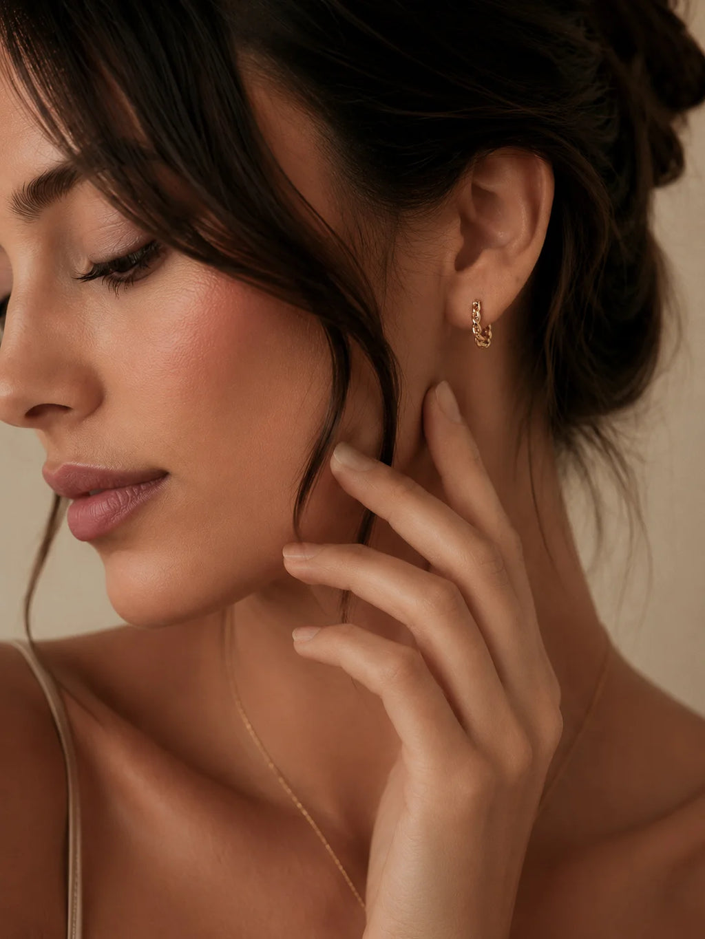A close-up lifestyle shot of a woman wearing a delicate gold huggie hoop earring and a fine gold chain necklace, set against a warm beige background.