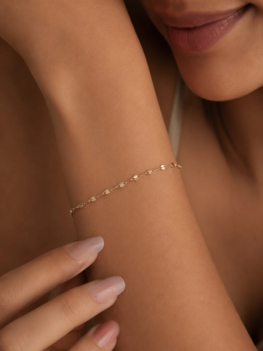 A delicate gold chain bracelet with small, repeating heart-shaped links, shown worn on a woman's wrist. The close-up shot features her hand resting near her wrist, with her lips and neck visible in the background, set against a warm, neutral skin tone.