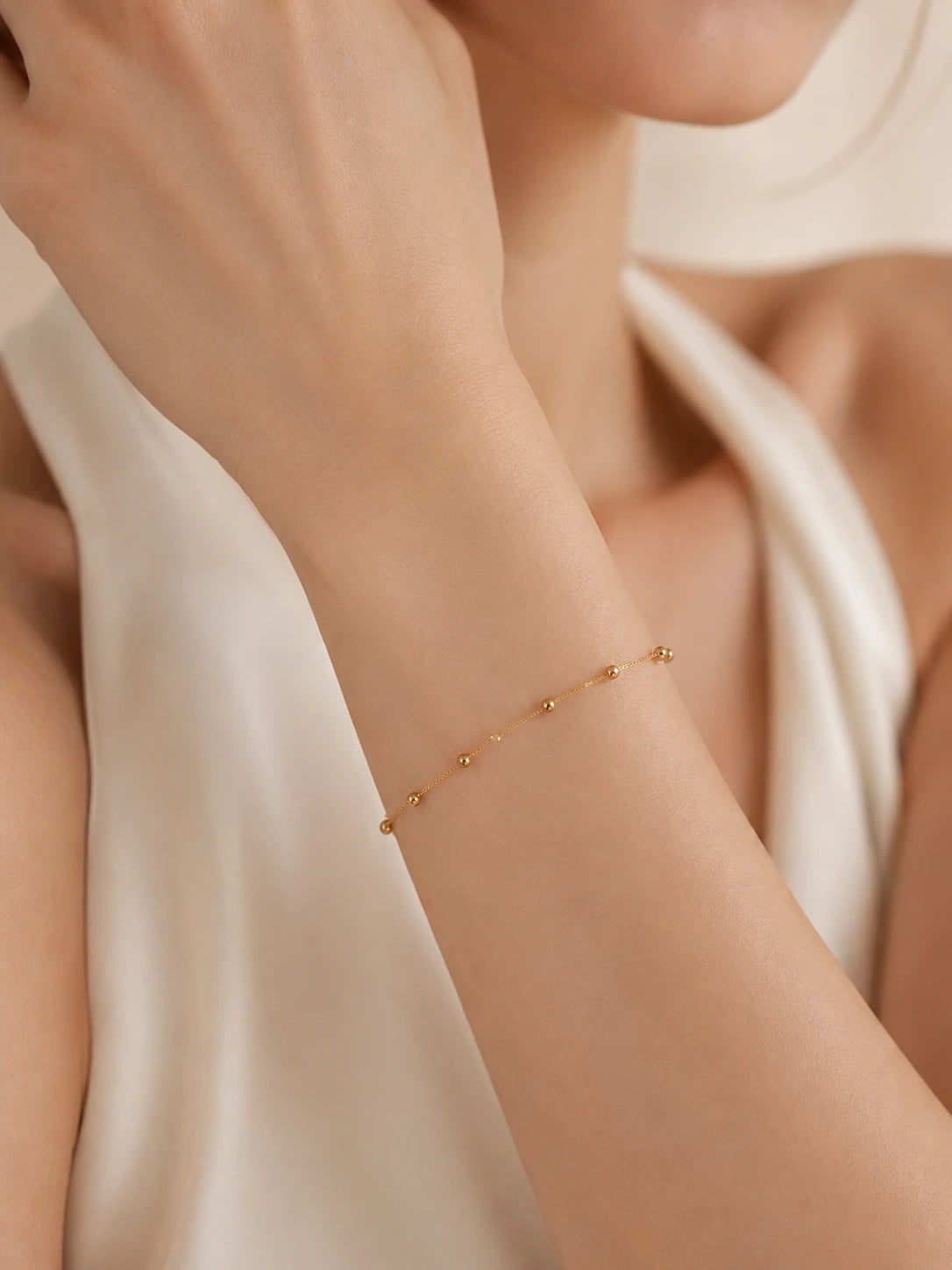 Close-up of 18K solid gold satellite bead chain bracelet on wrist, evenly spaced gold ball stations, fine gold chain, cream background