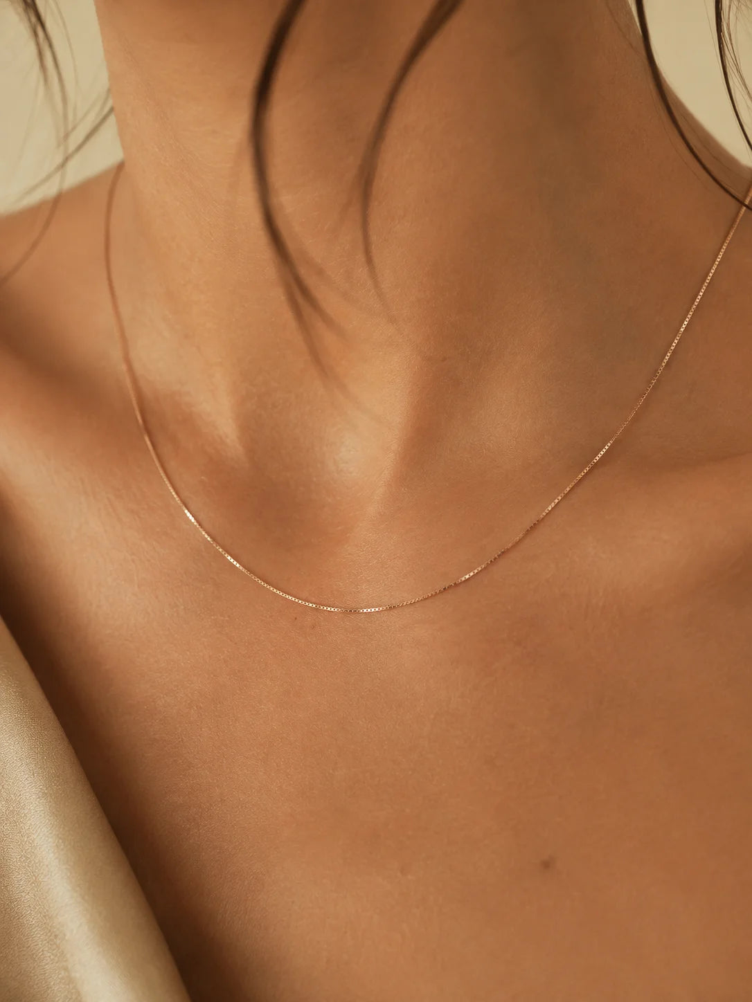 A delicate, fine chain necklace in a warm gold or rose-gold tone worn on a model's neck, shown in a close-up shot with a warm beige background.