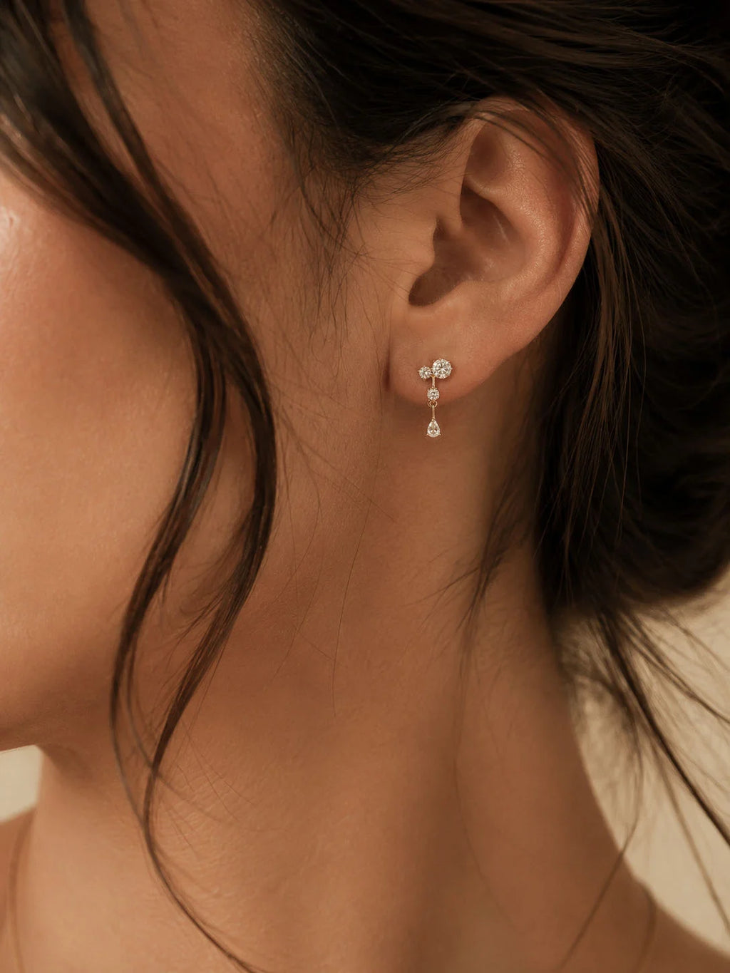 A close-up of a woman's ear wearing a delicate silver stud earring with a small cluster of clear stones and a dangling teardrop charm, set against warm skin tones and dark hair.