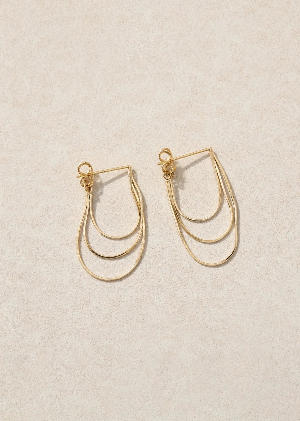 A pair of gold-toned, multi-layered hoop earrings with small decorative beads at the top, displayed on a textured beige background.