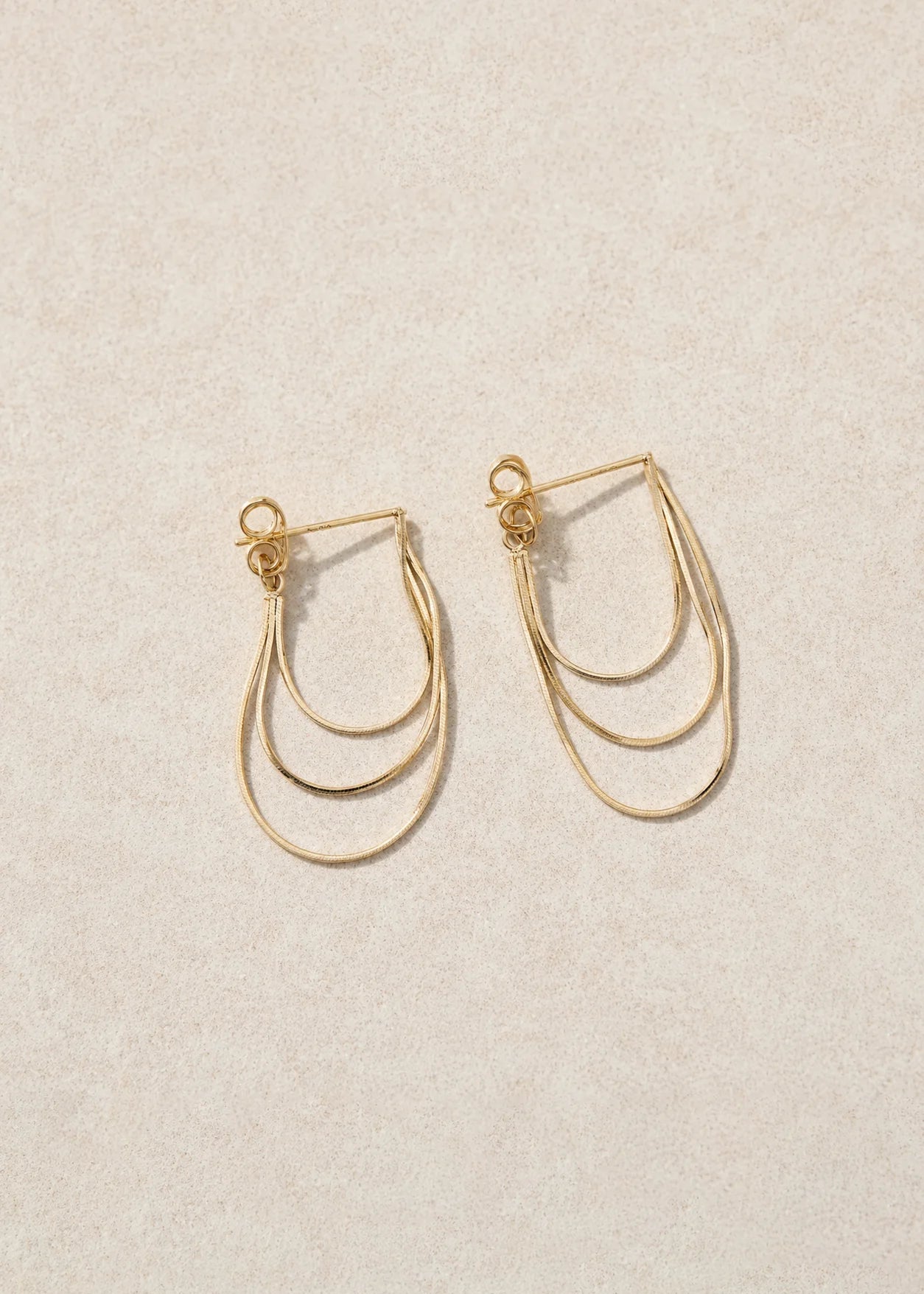 A pair of gold-toned, multi-layered hoop earrings with small decorative beads at the top, displayed on a textured beige background.