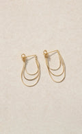 A pair of delicate gold-tone geometric hoop earrings with multiple curved bands, displayed on a textured beige background.
