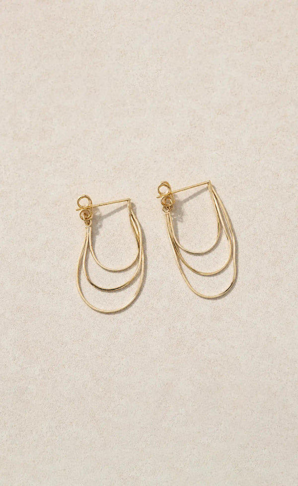A pair of delicate gold-tone geometric hoop earrings with multiple curved bands, displayed on a textured beige background.