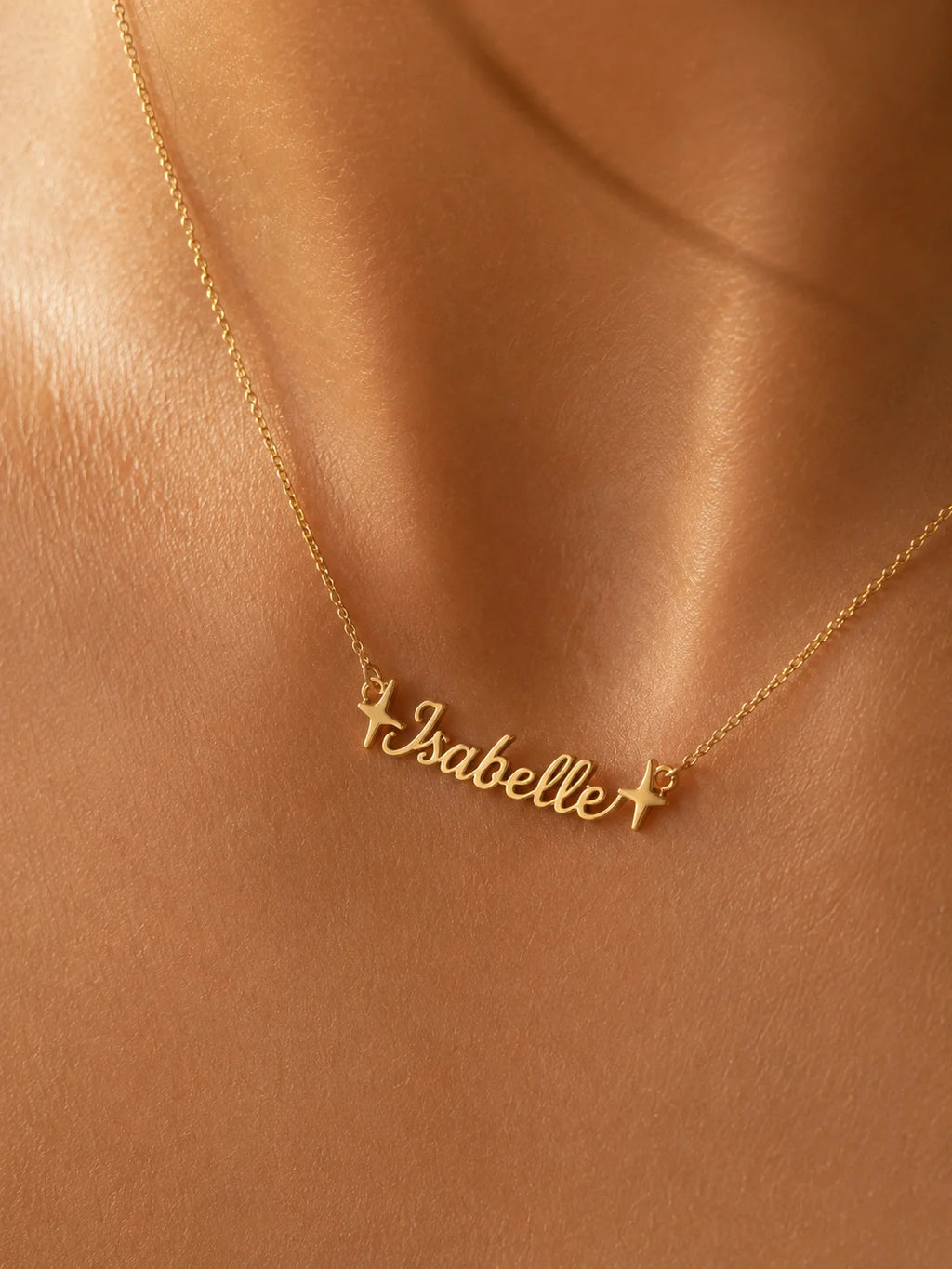 A gold personalized necklace with the name 'Isabelle' in cursive script, flanked by two small stars, worn on a model's neck against a warm, satin-like background.