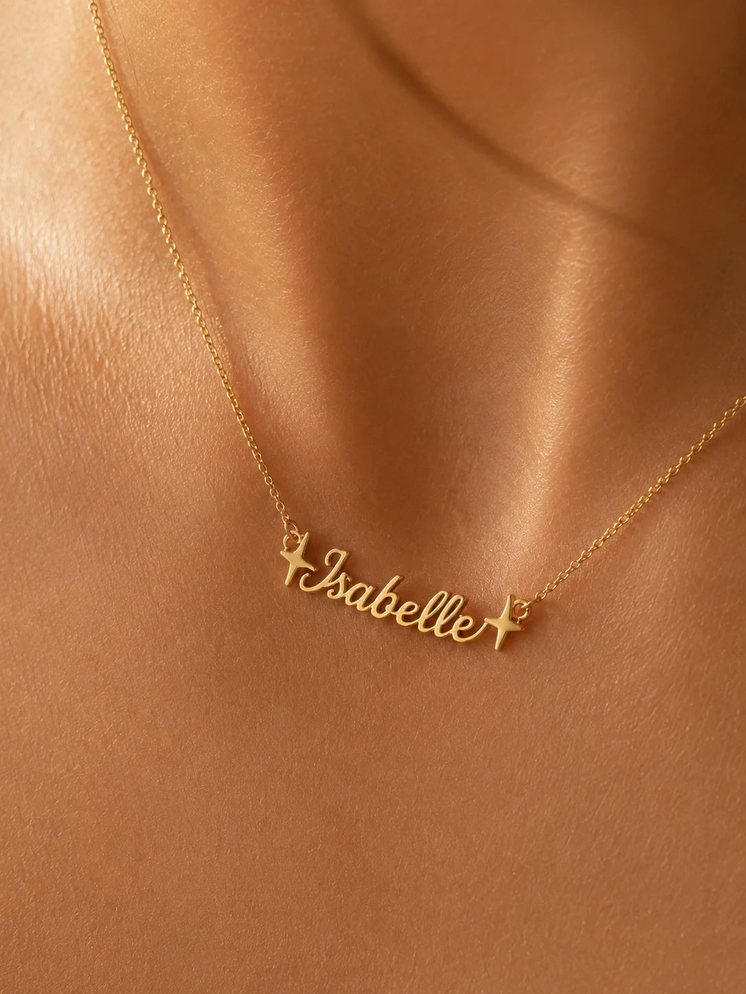 A gold personalized necklace with the name 'Isabelle' in cursive script, flanked by two small stars, worn on a model's neck against a warm, satin-like background.