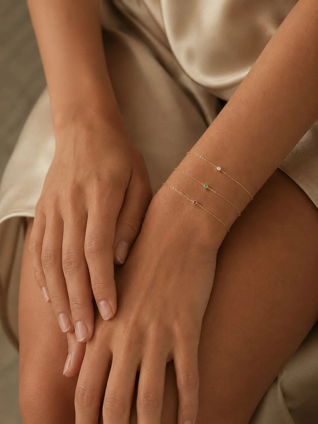 A close-up of a person's hands and wrist wearing three delicate gold chain bracelets, each adorned with a small colorful gemstone, set against a soft beige satin garment.