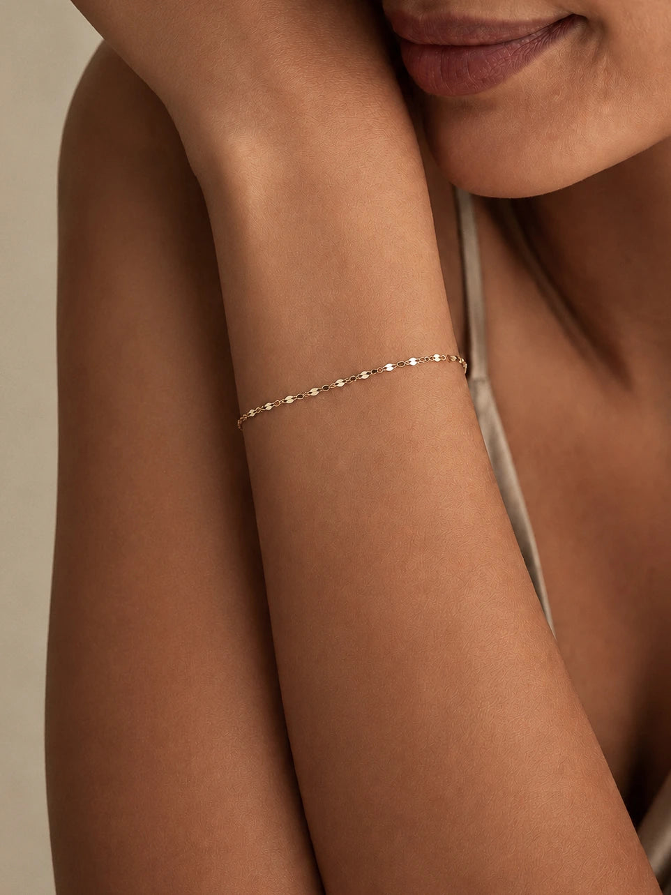 A delicate gold chain bracelet worn on a woman's wrist, shown in a close-up lifestyle shot against a warm beige background.