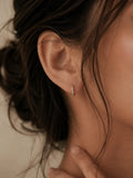 A close-up of a model's ear wearing a delicate gold huggie hoop earring with small clear stones, set against a soft beige background.