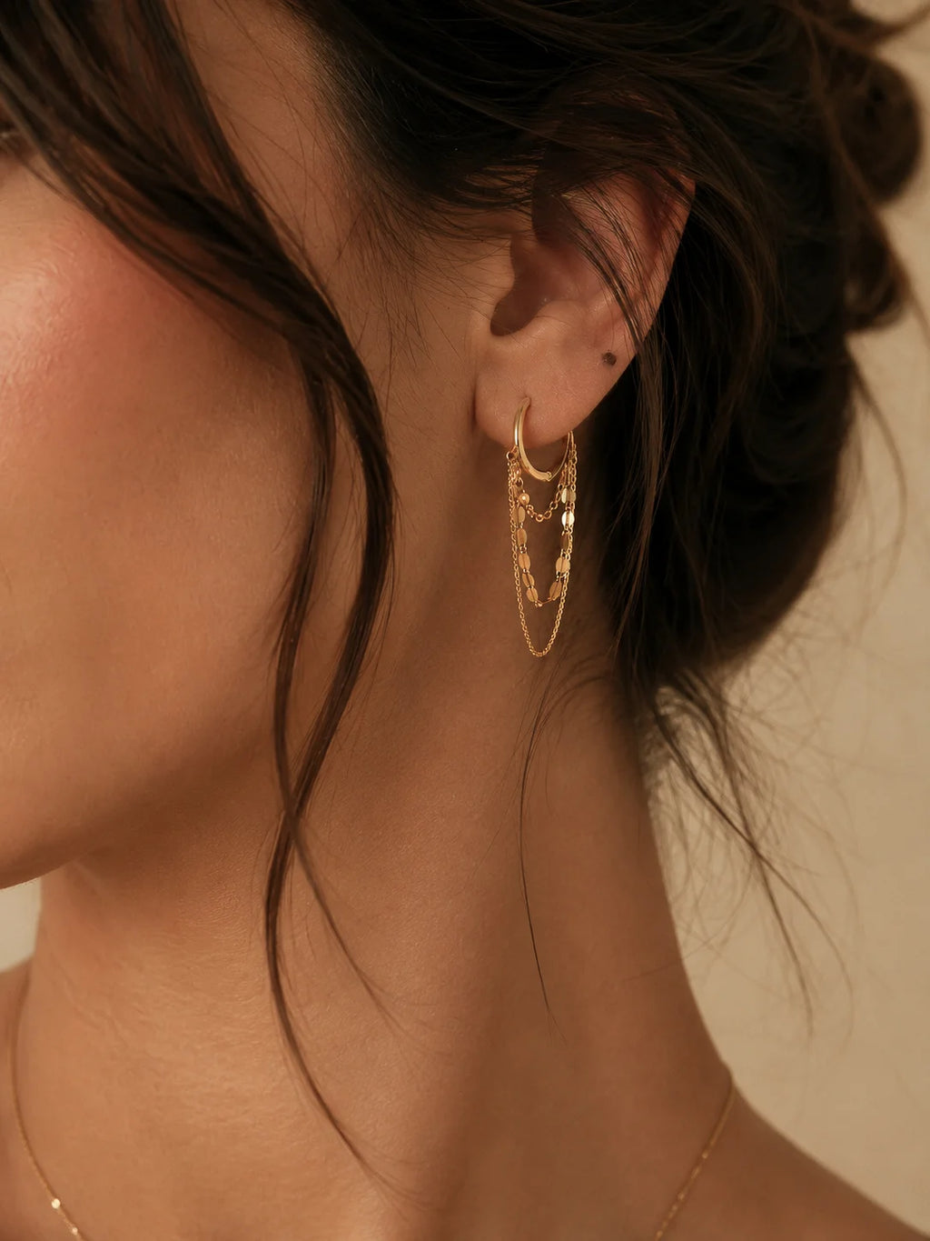 A close-up of a model's ear wearing a pair of gold hoop earrings with textured details, set against a warm beige background.