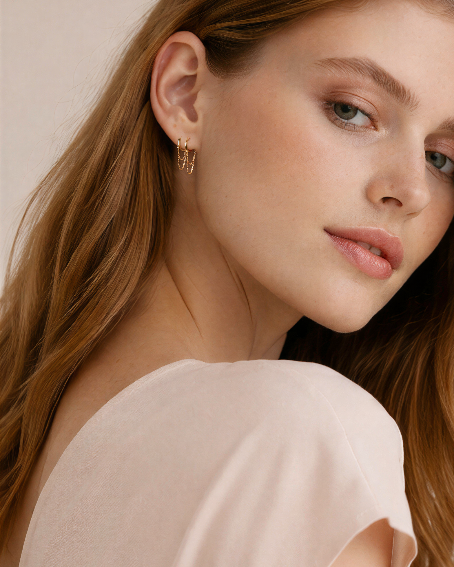 Woman wear double chain hoop with long brown hair wearing a light pink top against a neutral background