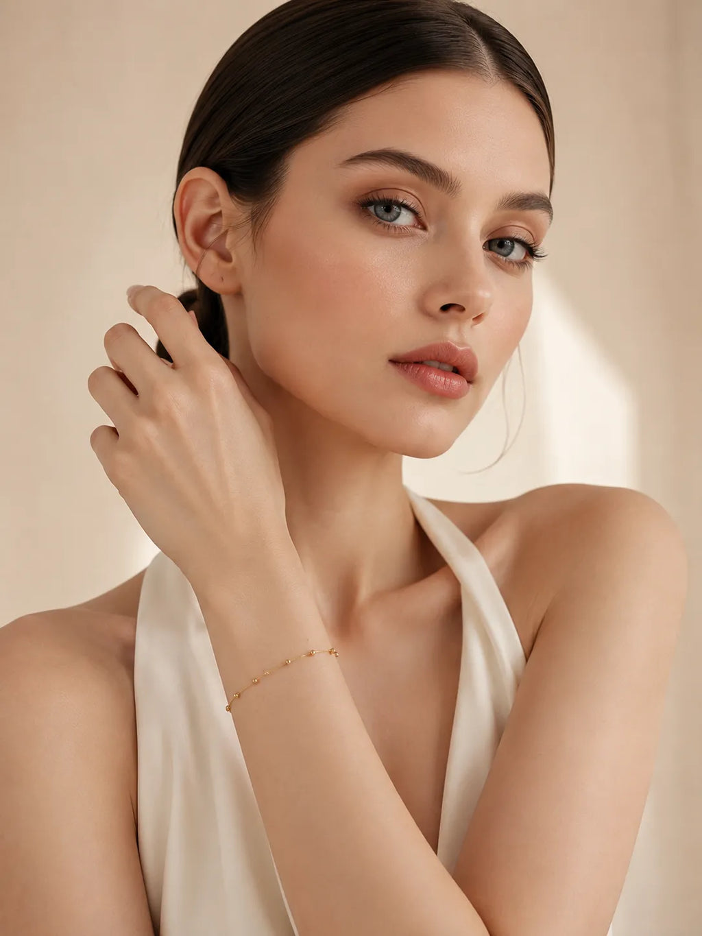 18K solid gold bead station bracelet on woman's wrist, model with hand raised to neck, styled with white satin halter top, warm beige background