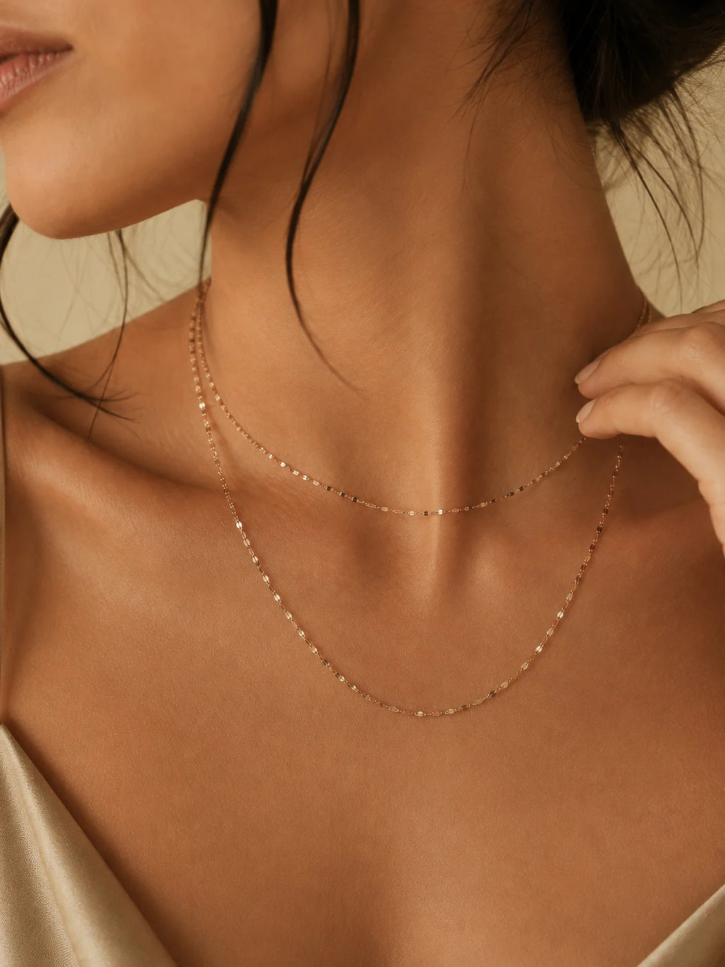 A close-up lifestyle shot of a model wearing a delicate double-layered rose gold chain necklace. The necklace features tiny, evenly spaced beads on both layers, styled with a satin top against a warm beige background.