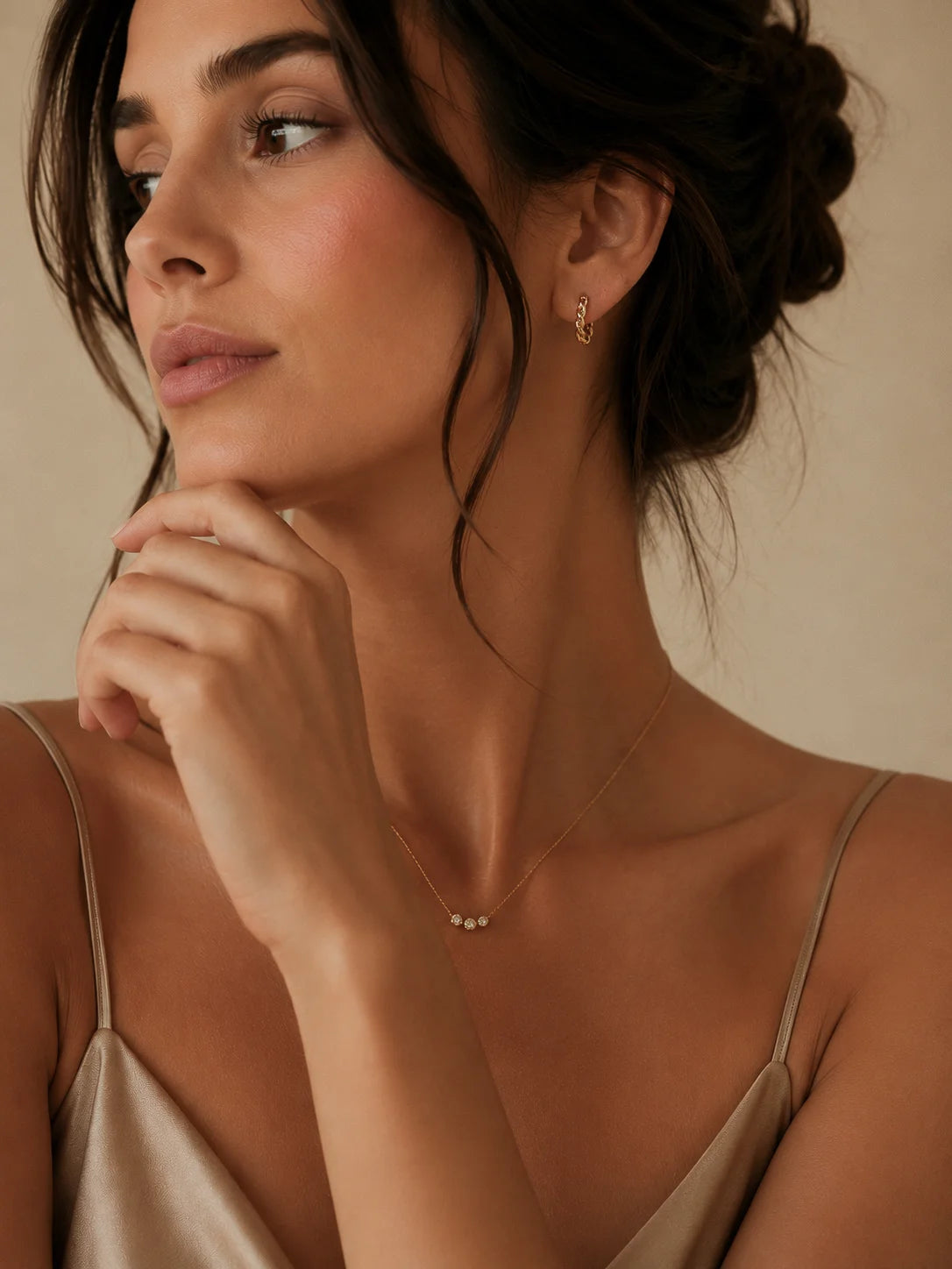 A close-up lifestyle shot of a woman with dark hair in an updo, wearing a delicate gold necklace with three small pearls and a small gold hoop earring, set against a warm beige background.